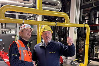 A photo of a STRABAG employee and an ELCO employee are having a discussion in a building’s technical room. Both are wearing safety helmets.